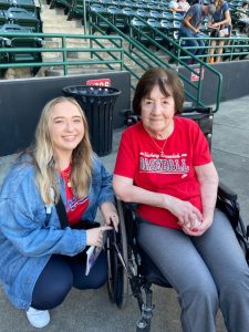 young woman with blonde hair kneels beside senior woman in wheelchair at hickory crawdads baseball game