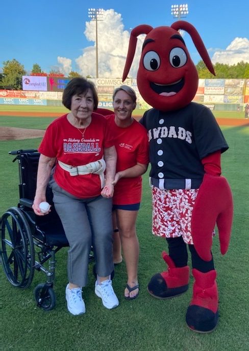 Senior woman standing up from wheelchair with young woman supporting her and Hickory Crawdads mascot Conrad the Crawdad