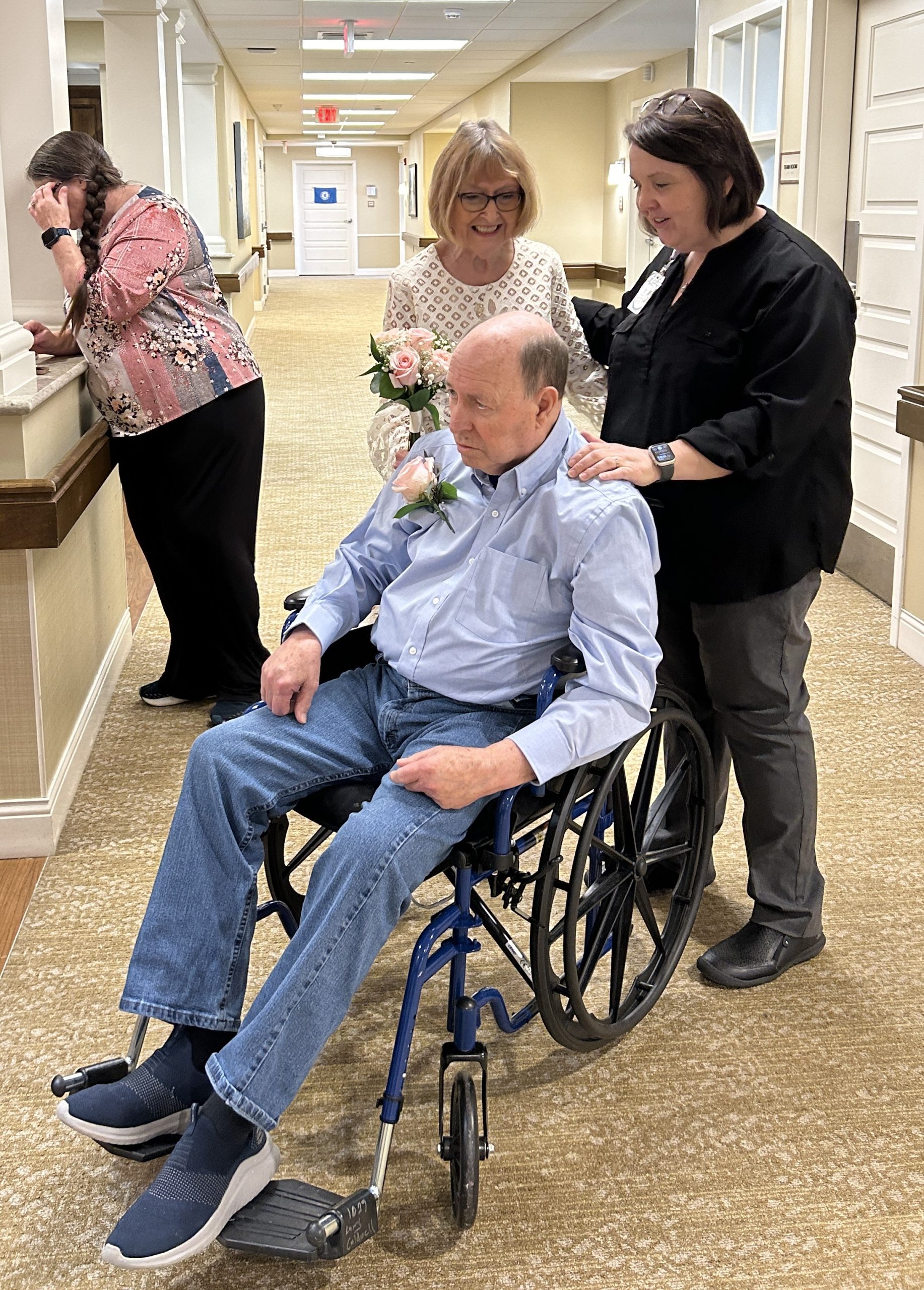 senior woman in white dress with bouquet stands beside man in wheelchair and beside dark-haired woman with her hand on his shoulder